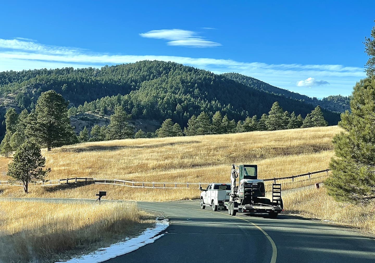 A Liftech truck towing a Bobcat on a trailer along a Front Range road, with foothills and golden grass